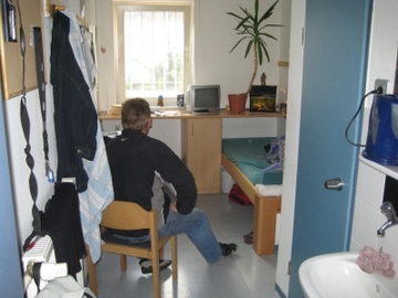 Foto eines Einzelhaftraumes in der sozialtherapeutischen Abteilung. Ein Gefangener sitzt auf einem Stuhl mit dem Rücken zur Kamera, umgeben von einem Bett, Tisch Stuhl, Regal, Wasserkocher und diversen anderen Gegenständen (Zum Artikel Sozialtherapie).