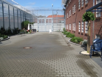 Foto des Freistundenhofes, links ist ein hoher Zaun und rechts das Gebäude der Sozialtherapie abgebildet (Zum Artikel über die Sozialtherapie). Suchsymbol Lupe