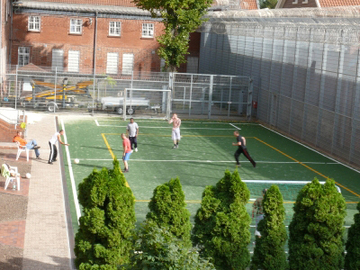 Das Foto zeigt ein Kleinspielfeld aus der Vogelperspektive auf dem Fußball gespielt wird (Zum Artikel Sport und Freizeitangebote in der Hauptanstalt). Suchsymbol Lupe
